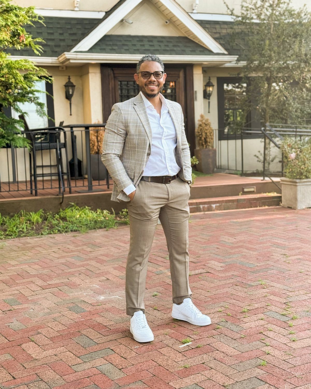 Man wearing a patterned blazer, tan pants, and white sneakers on a brick patio.