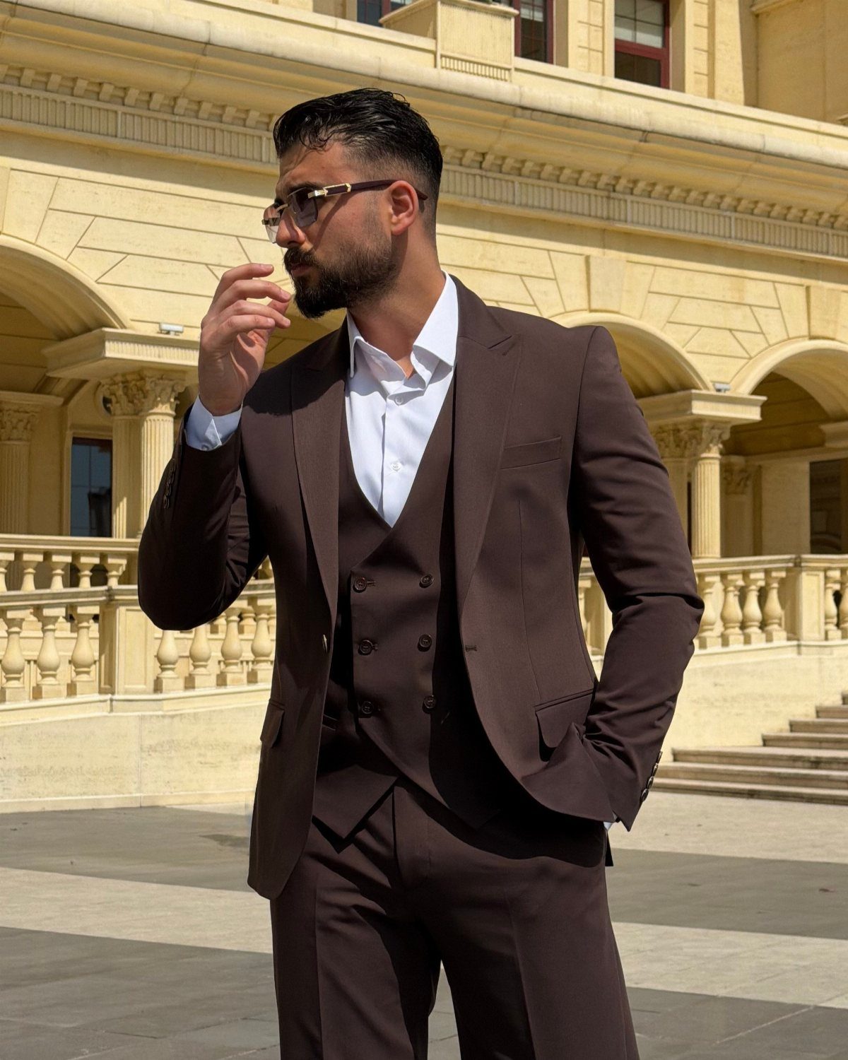 Man in a dark brown three-piece tailored suit standing before a classical stone building.