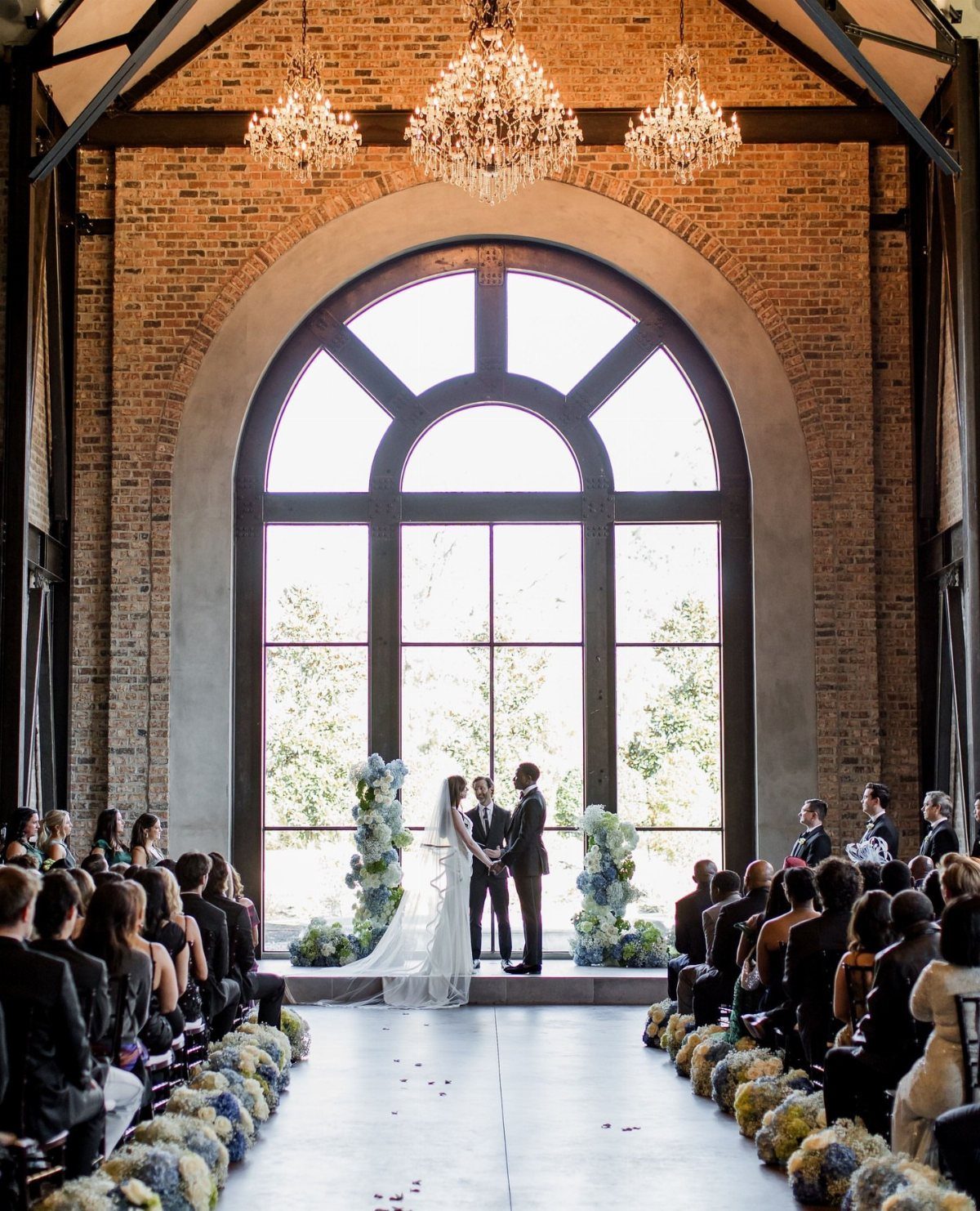 Wedding ceremony in a brick venue with a large arched window and chandeliers.