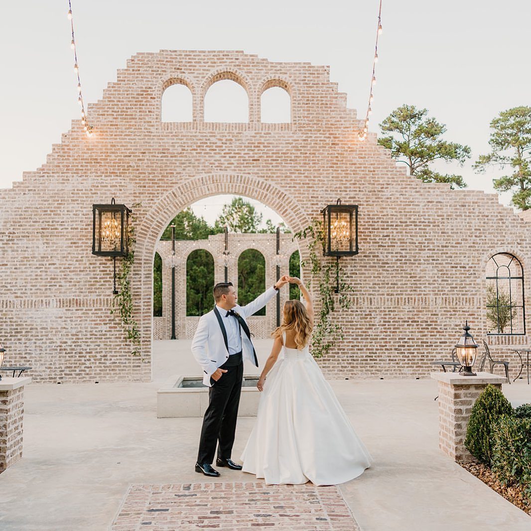Bride and groom dancing before a large brick wall with arched openings and lanterns.