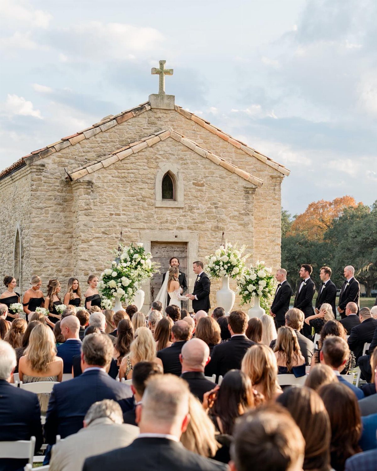 Wedding ceremony guests seated before a stone chapel with white floral arrangements.