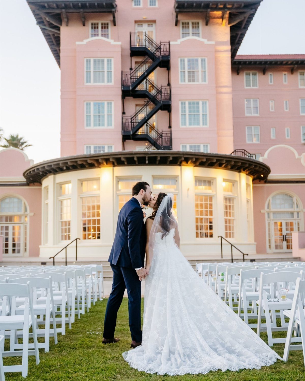 Bride and groom kissing before a pink Mediterranean building with white wedding chairs.