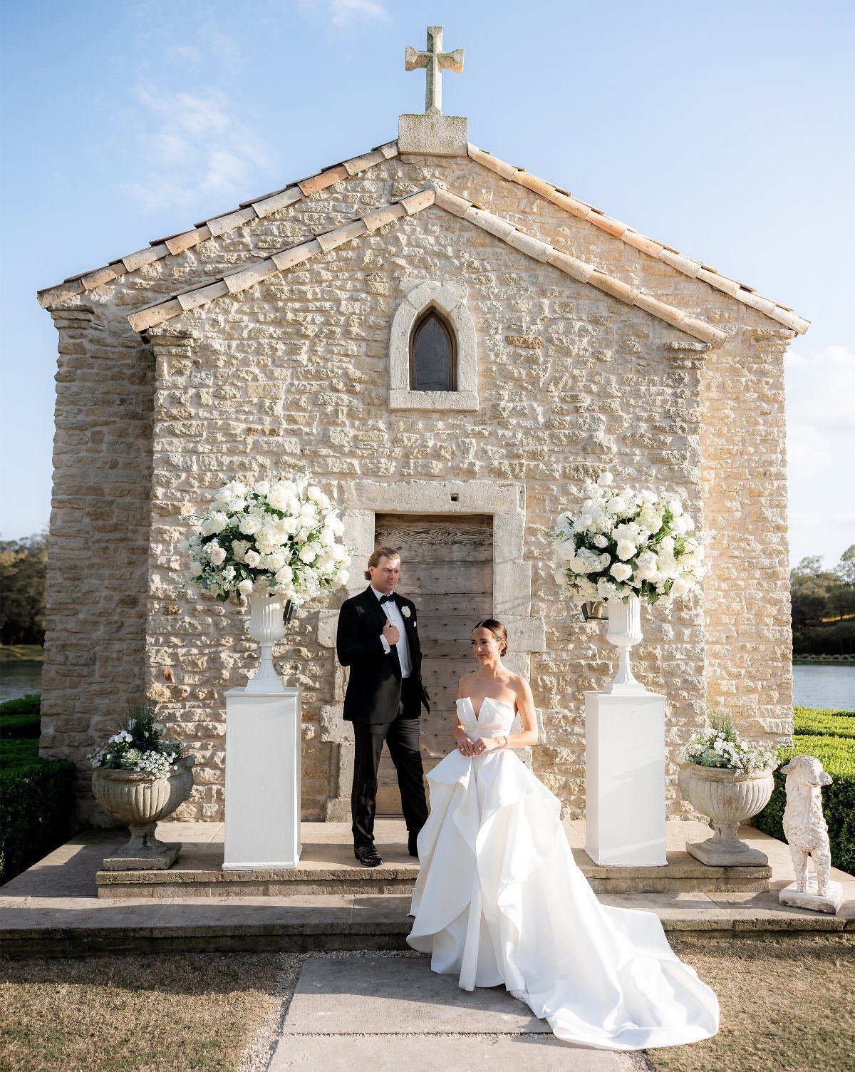 Bride and groom posing before a rustic stone chapel with white floral arrangements.