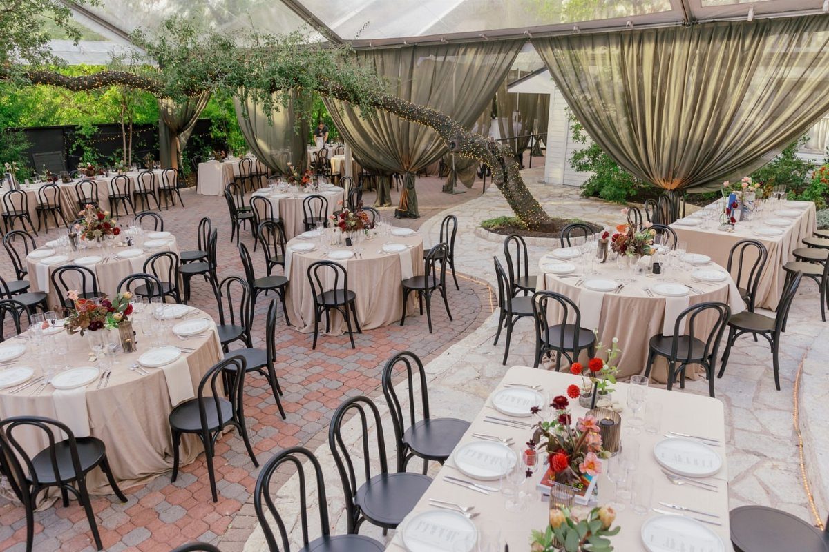 Courtyard wedding reception with beige linens, black chairs, and draped fabric under a canopy.