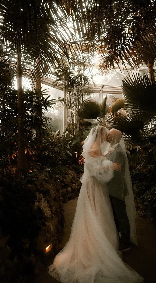 Bride and groom kissing in a lush indoor botanical garden conservatory with palms.