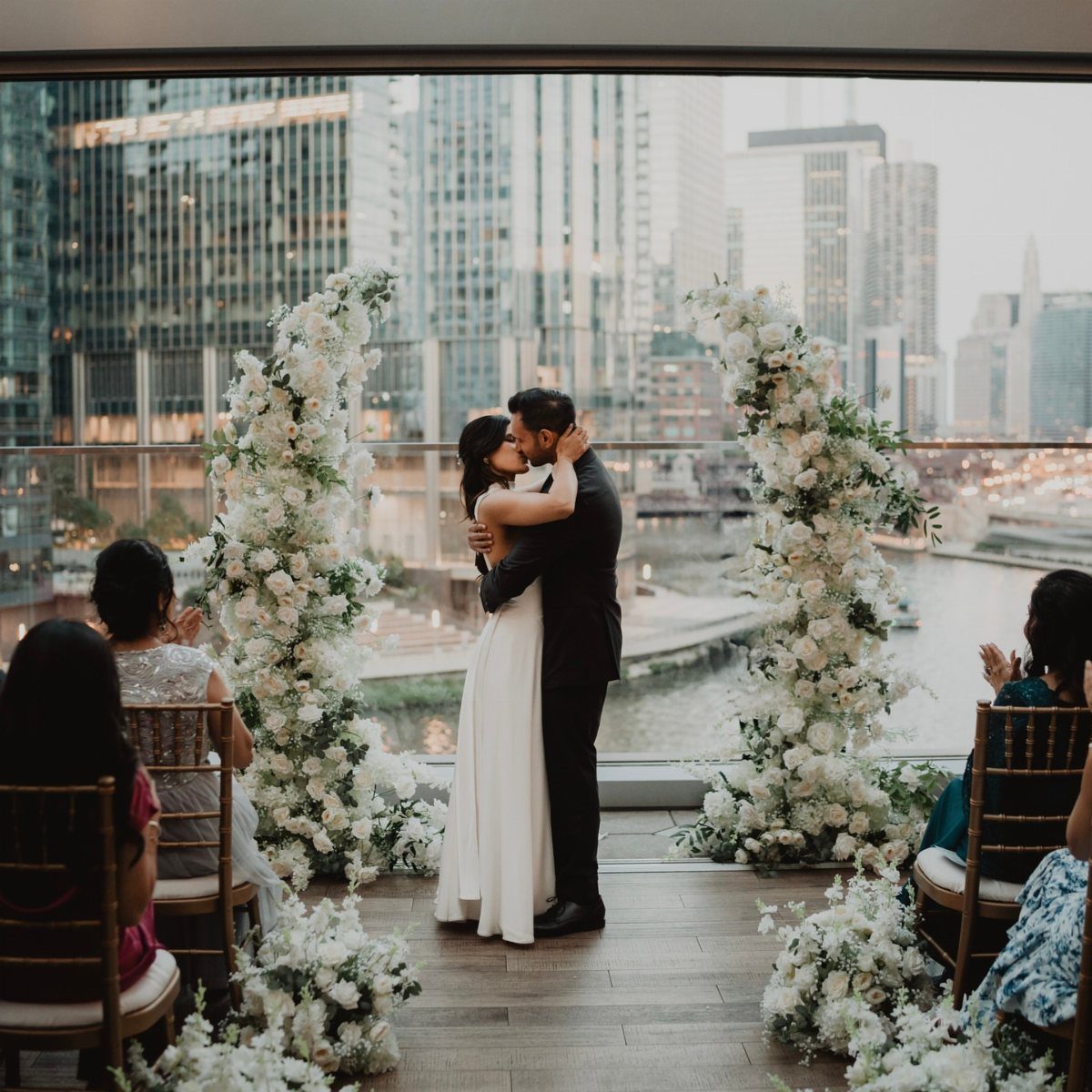 Couple kissing at a rooftop wedding ceremony with white floral arches and city skyline.