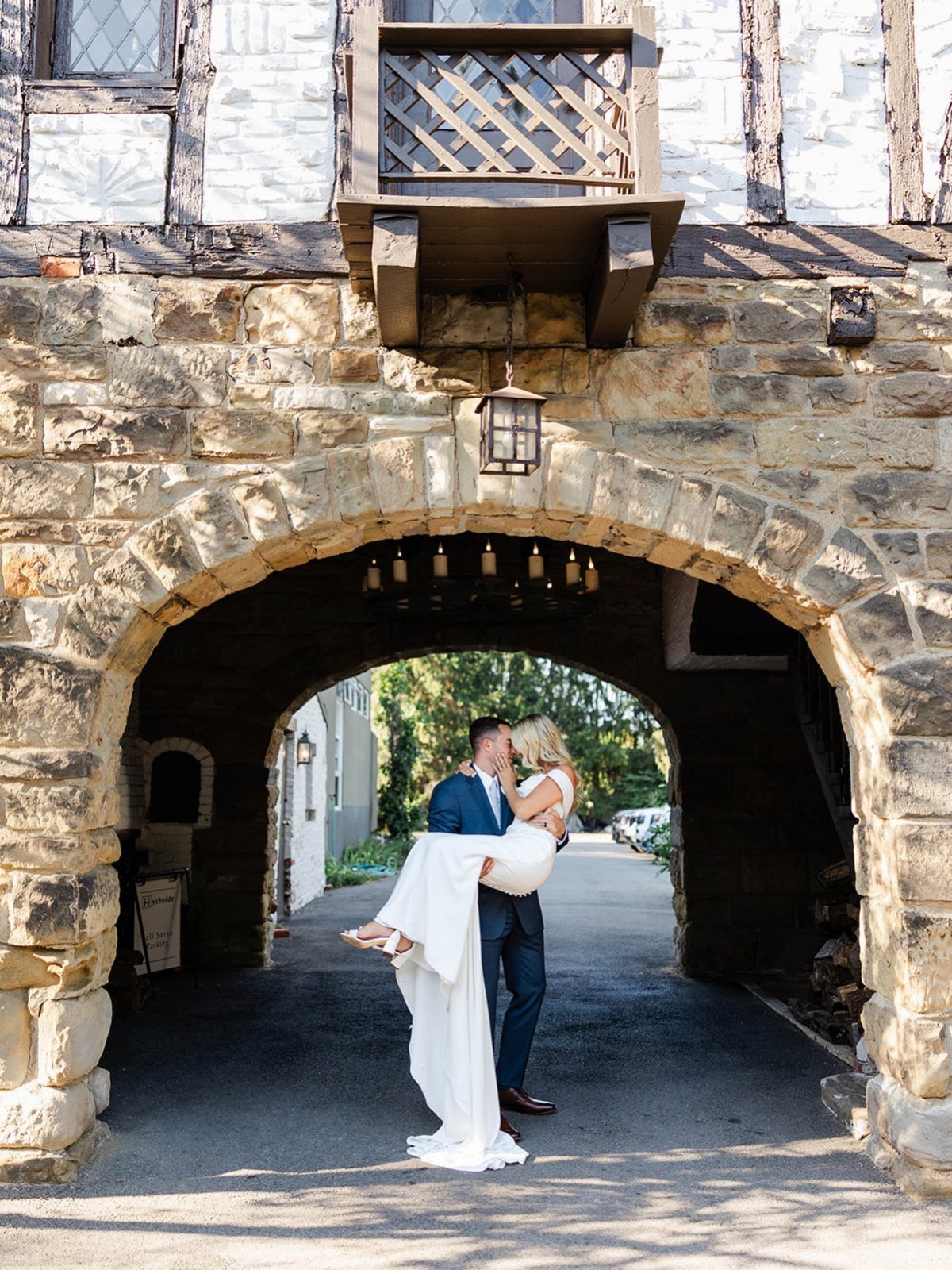 Groom carrying bride under a stone archway of a Tudor-style wedding venue.