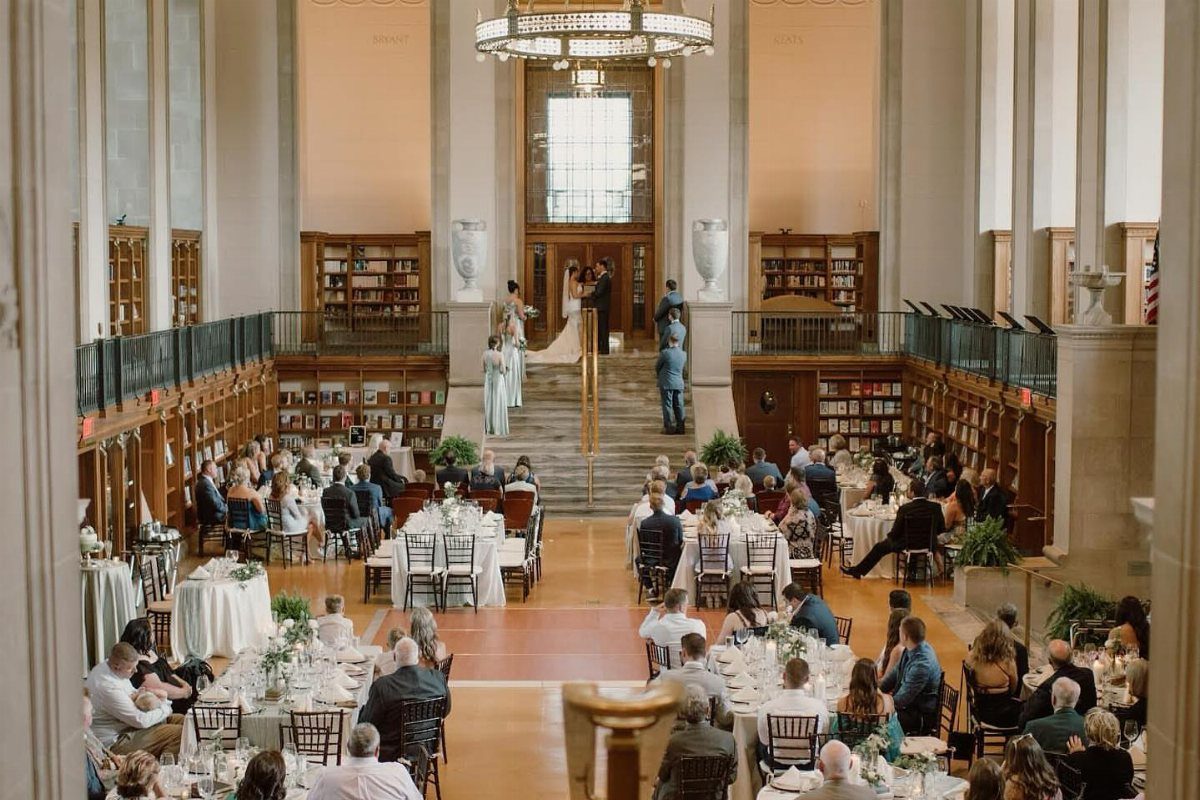 Wedding reception in a grand library with guests at tables and a couple on stairs.