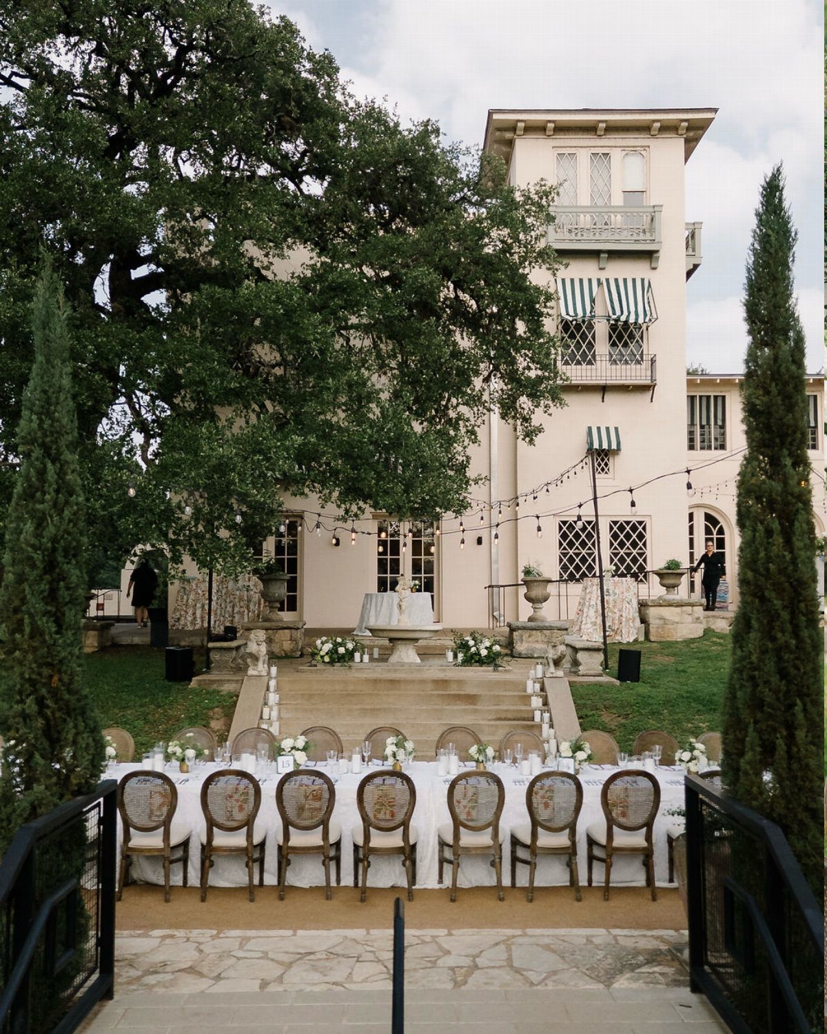Long white wedding table set before a cream Tuscan villa with tall cypress trees.