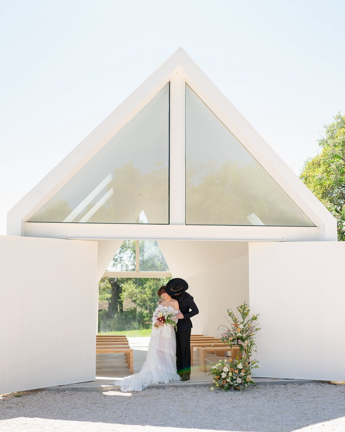 Bride and groom embracing in the doorway of a modern white A-frame chapel.