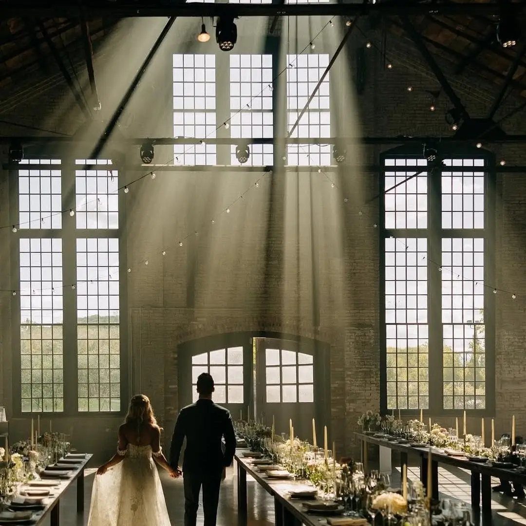 Bride and groom in an industrial wedding venue with long tables and sunbeams.