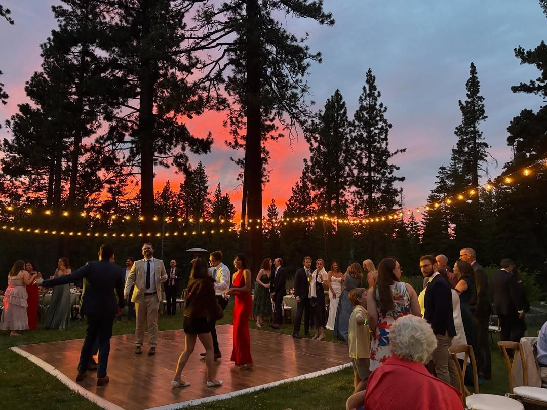 Wedding guests dancing on an outdoor wooden floor under string lights at sunset.