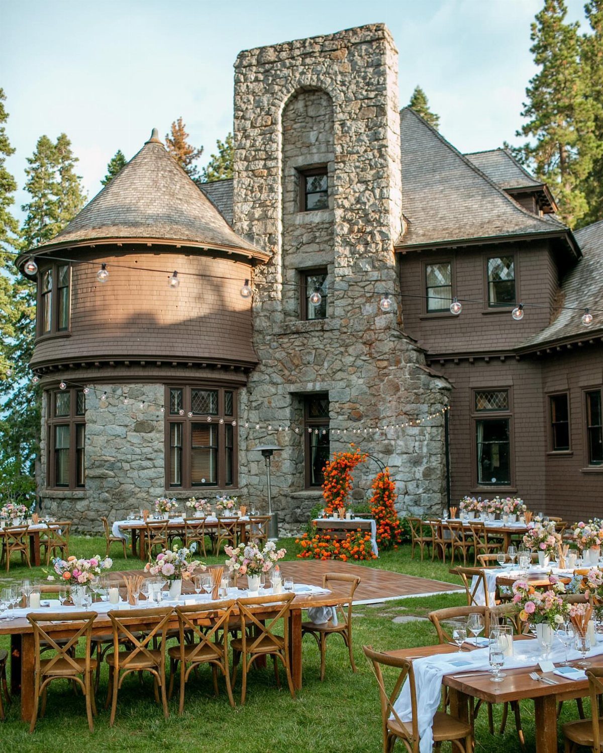 Outdoor wedding reception with wooden tables and floral arch before a stone estate.
