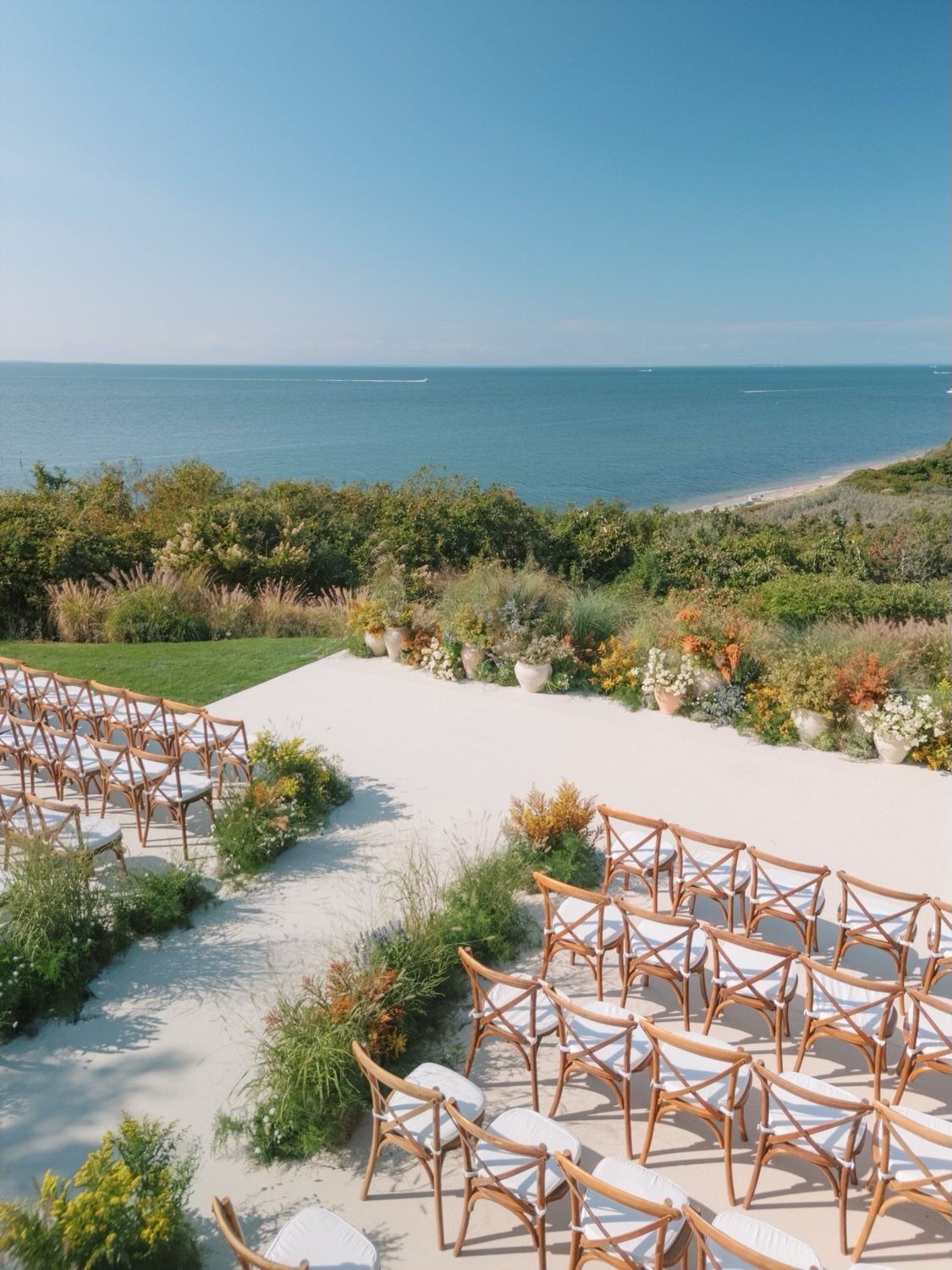 Outdoor wedding ceremony setup with wooden chairs on white sand overlooking the ocean.