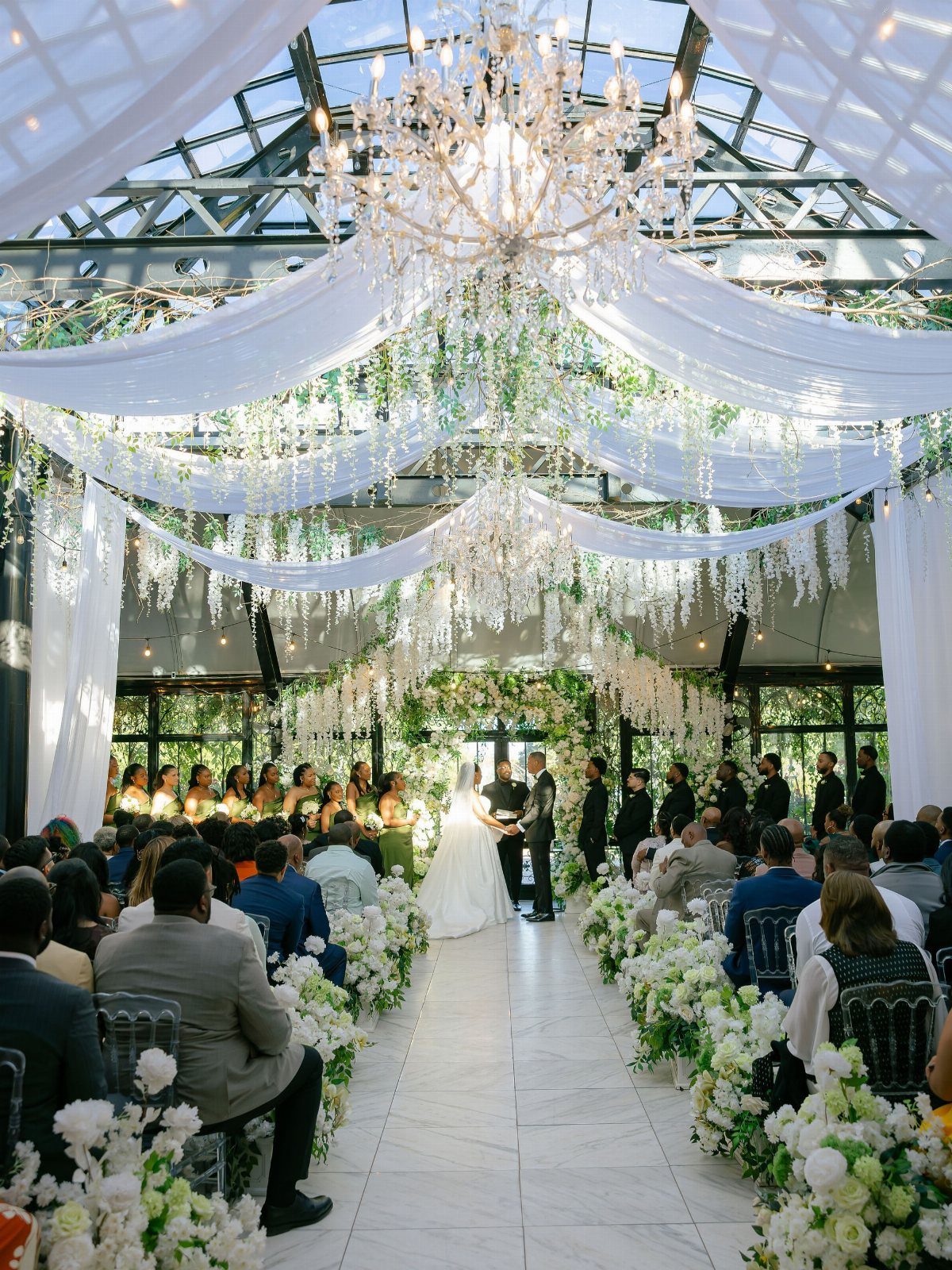 Wedding ceremony in a glass-roofed venue with white drapes, chandeliers, and floral aisle.