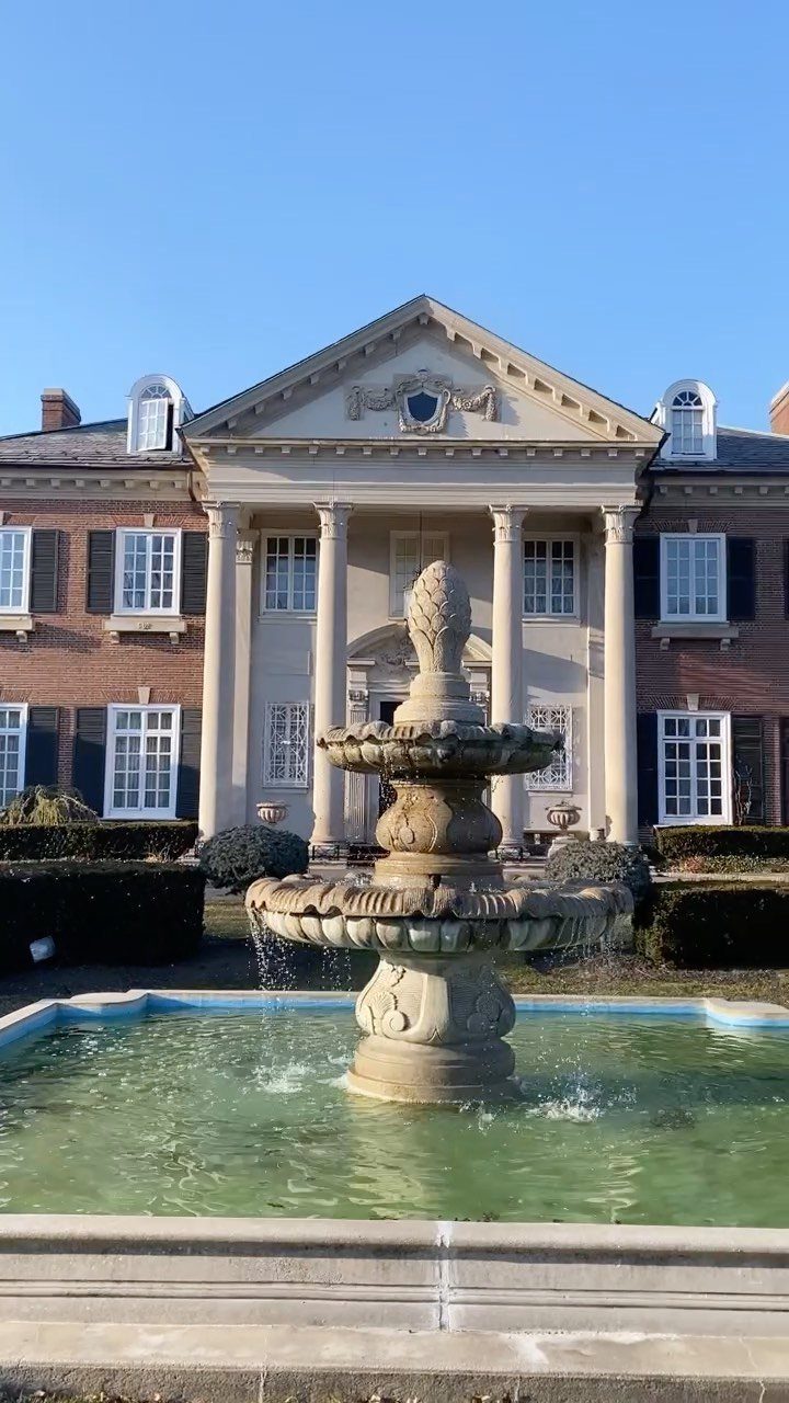 Tiered stone fountain in front of a grand red-brick mansion with white columns.