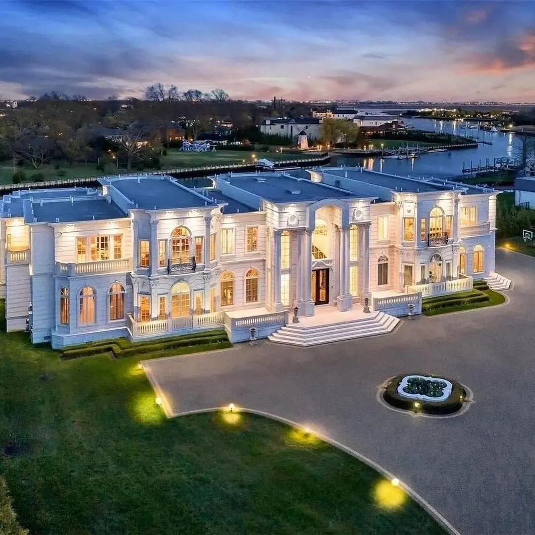Large white neoclassical mansion with illuminated windows and a circular driveway at dusk.
