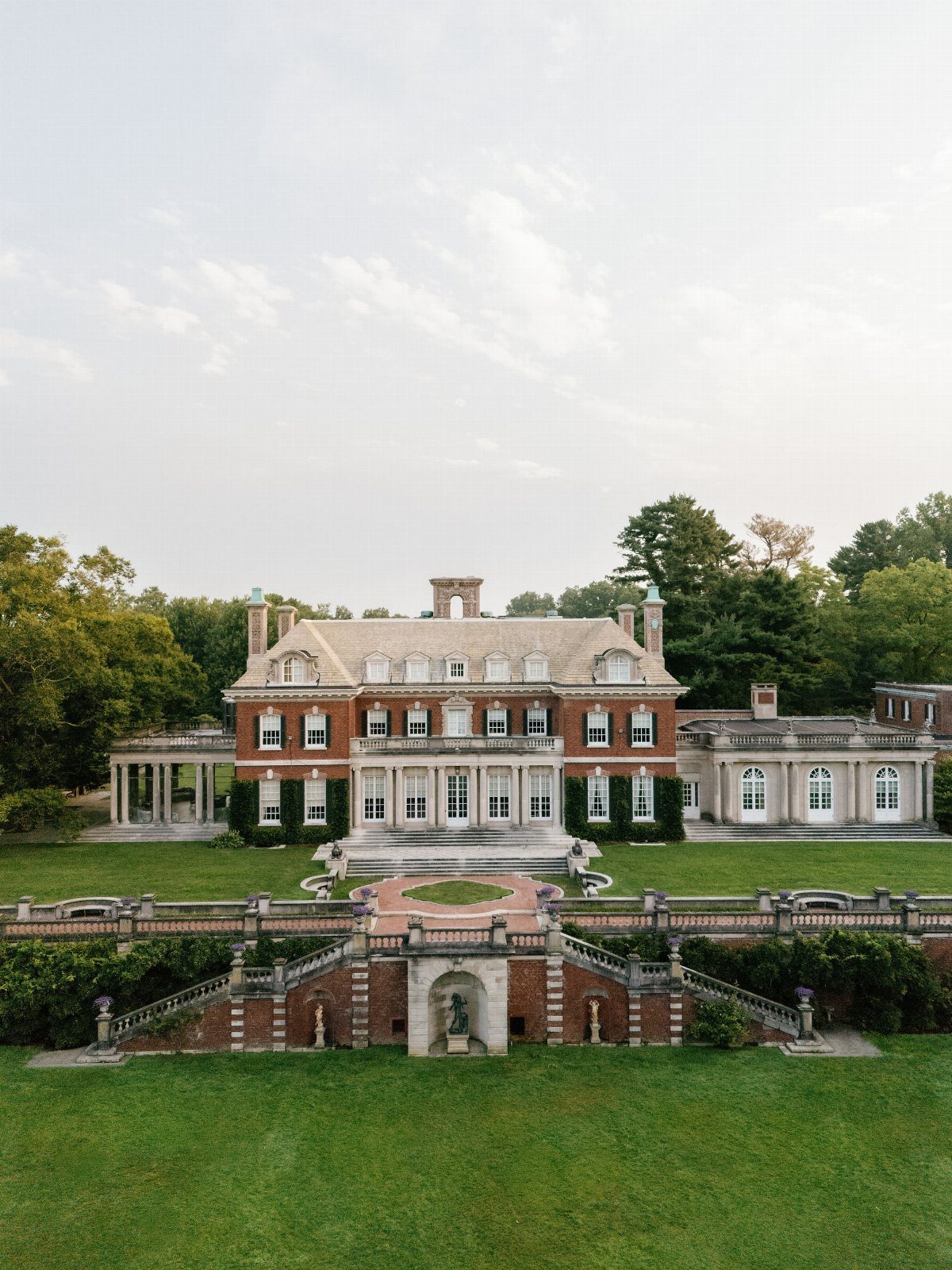 Historic red brick mansion with white columns and stone terraces facing a green lawn.