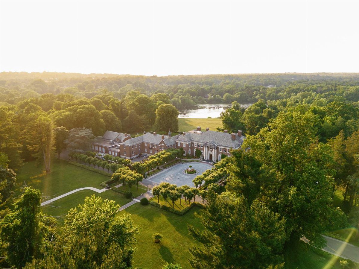 Large red-brick historic mansion with manicured gardens and a circular driveway in a forest.
