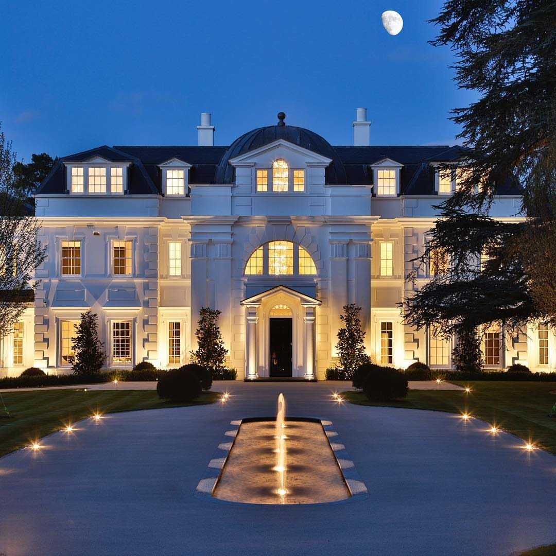 Large white luxury mansion illuminated at night with a reflecting pool and fountain.