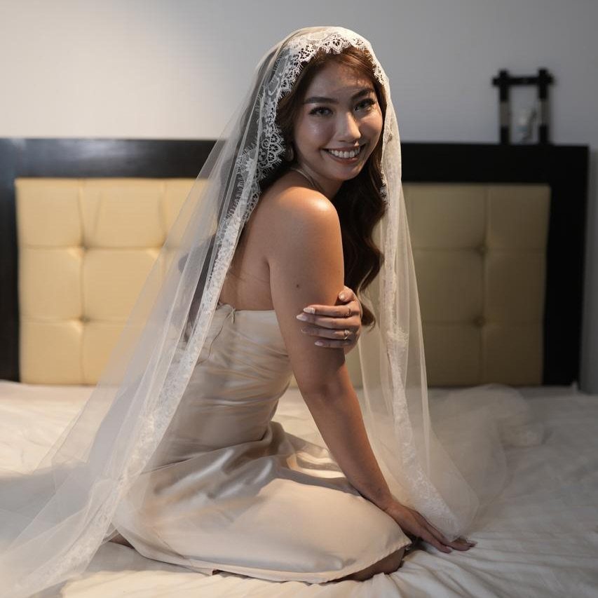Woman in a white satin dress wearing a lace mantilla wedding veil on a bed.