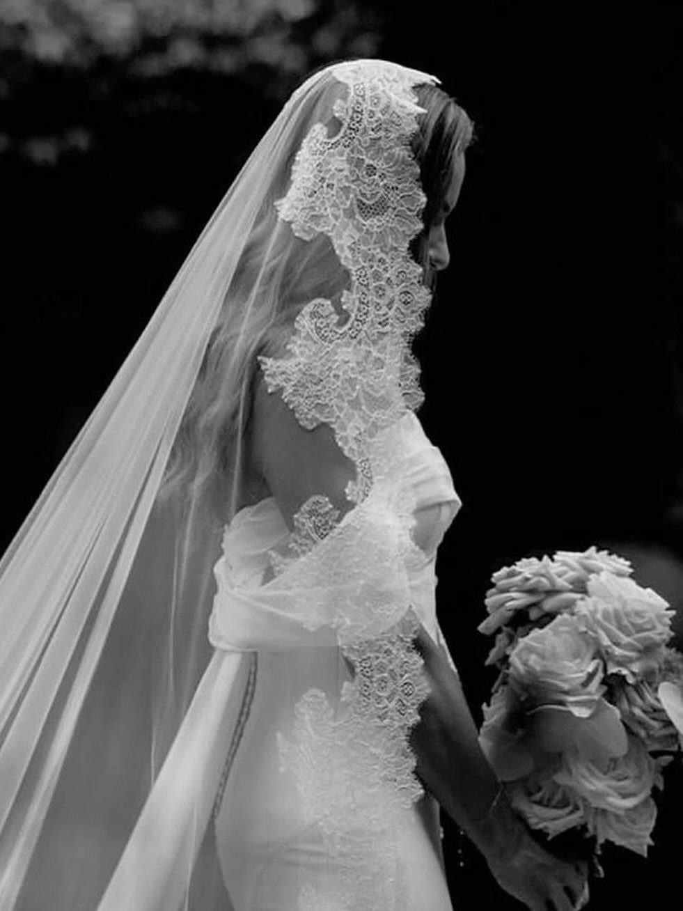 Bride in a white dress wearing a lace-trimmed mantilla wedding veil and holding roses.