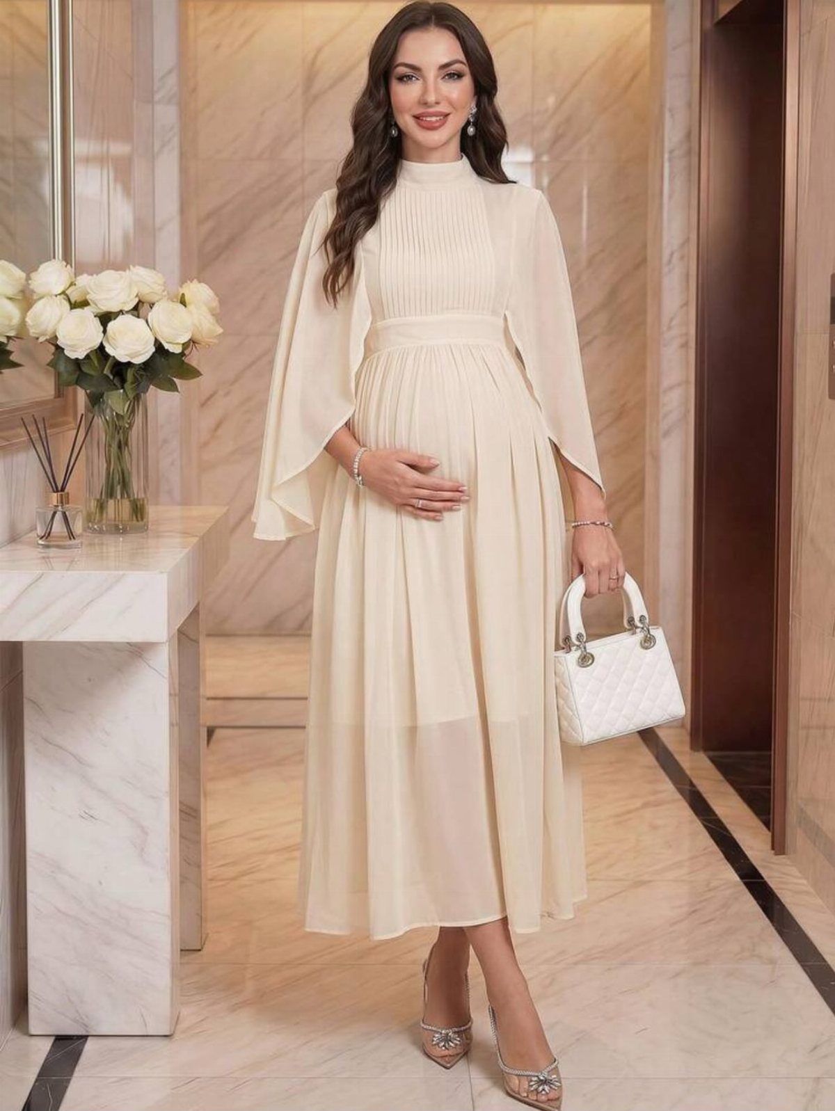 Pregnant woman in a cream high-neck maternity dress with cape sleeves and white handbag.