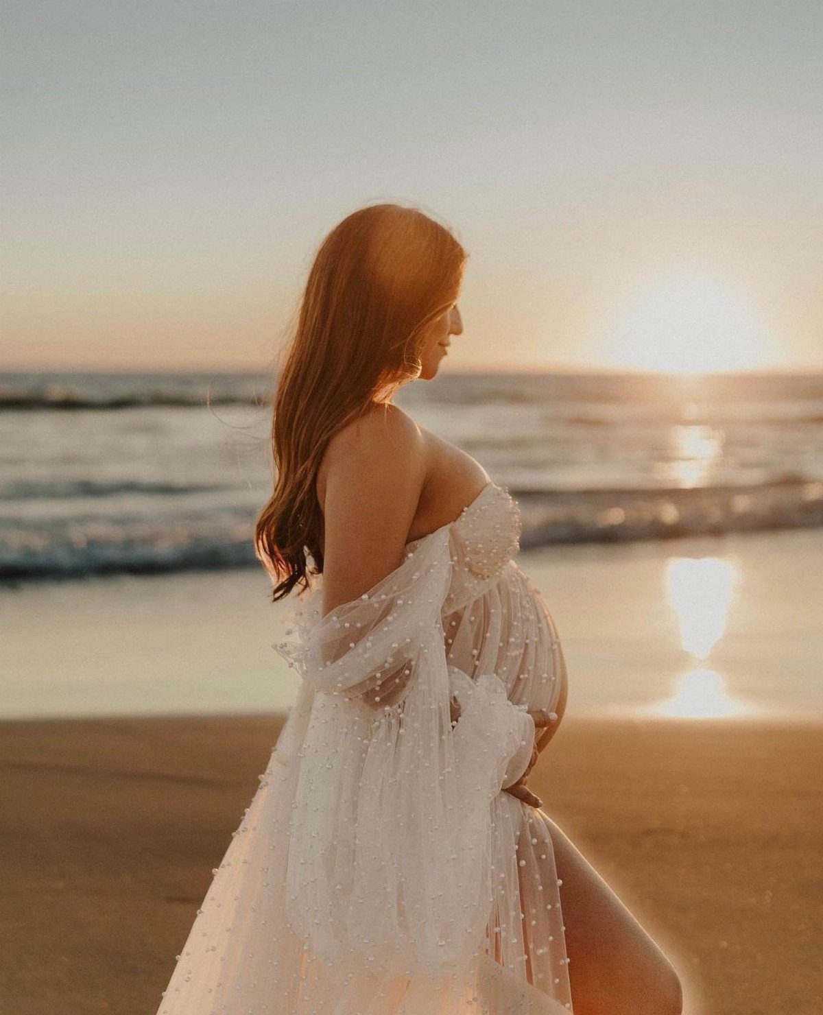 Pregnant woman in a white flowy maternity gown standing on a beach at sunset