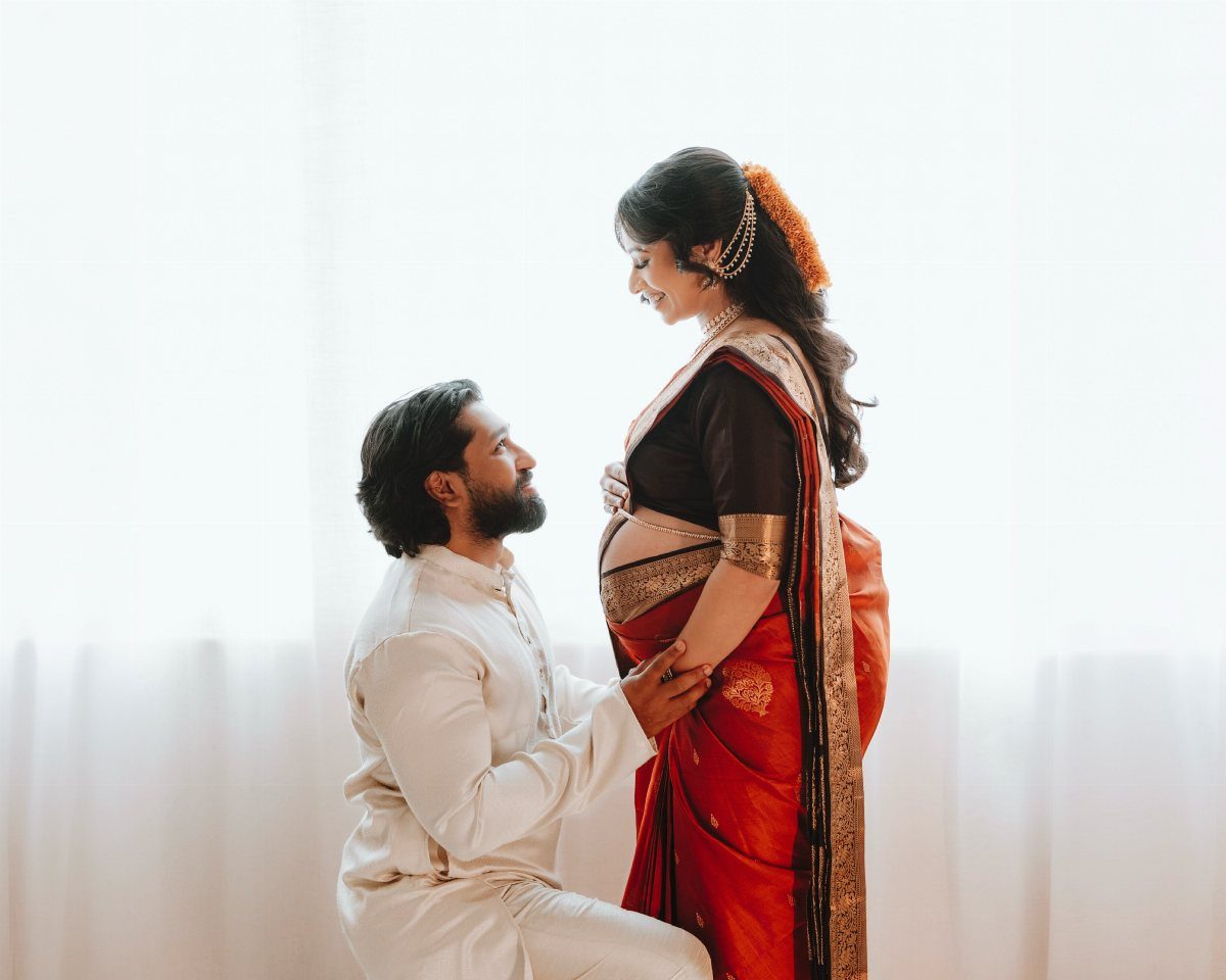 Pregnant woman in red saree and man in white kurta posing together.