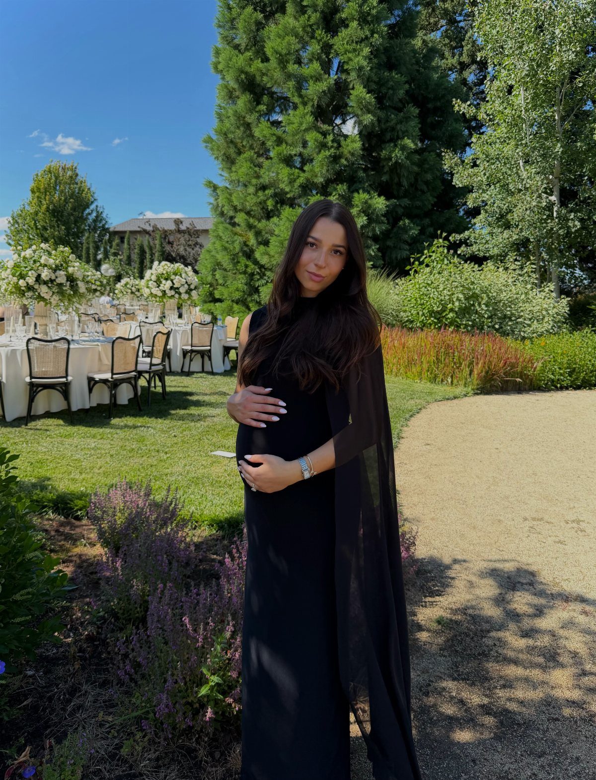 Pregnant woman in a long black dress and cape at an outdoor wedding.
