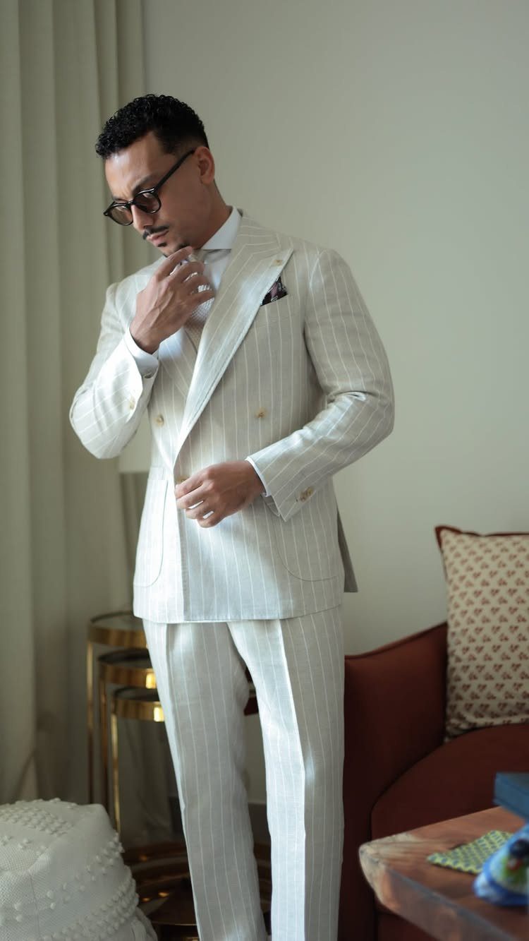 Man in a tailored light-colored pinstriped linen suit adjusting his tie indoors.