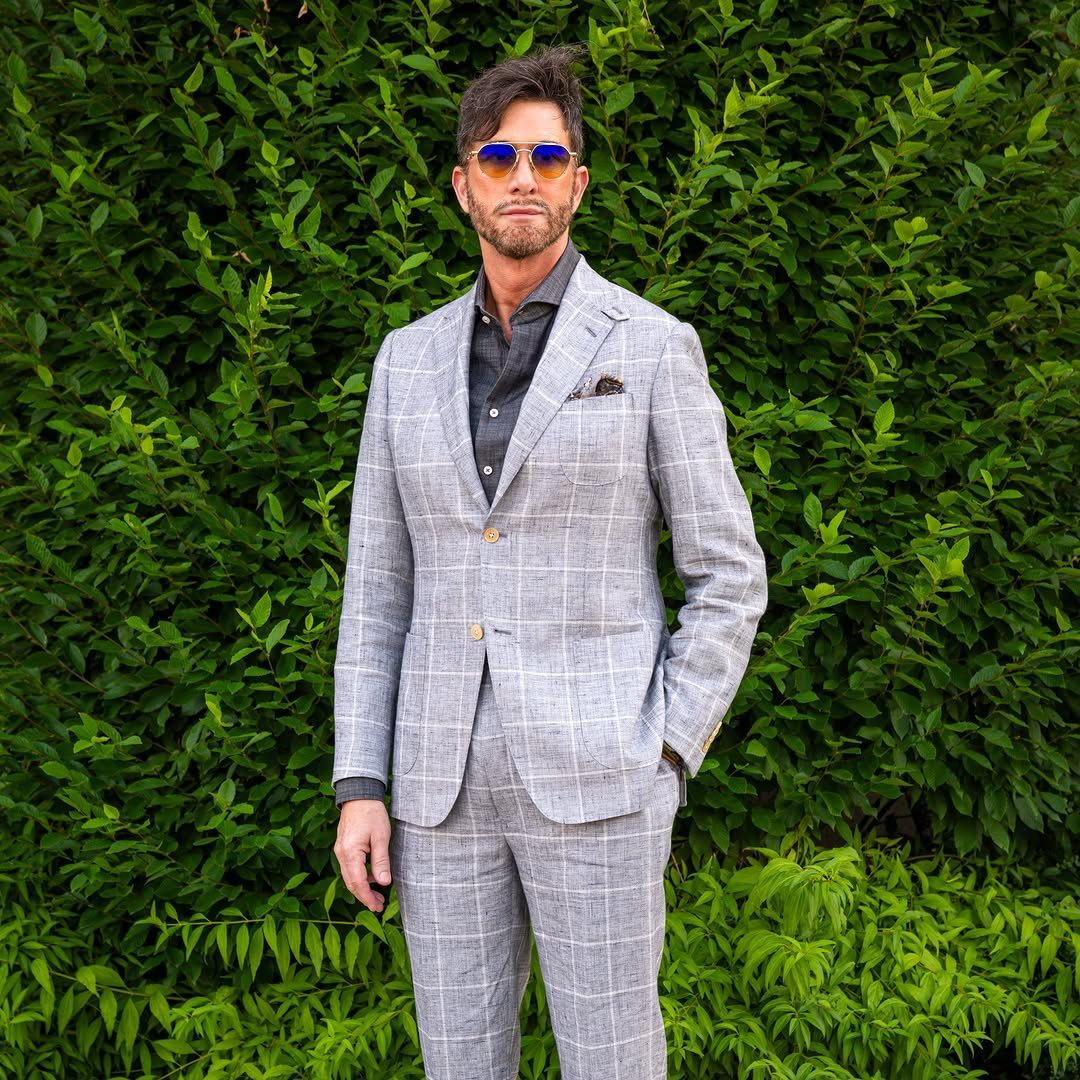 Man in a grey linen windowpane check suit and sunglasses against a green hedge.