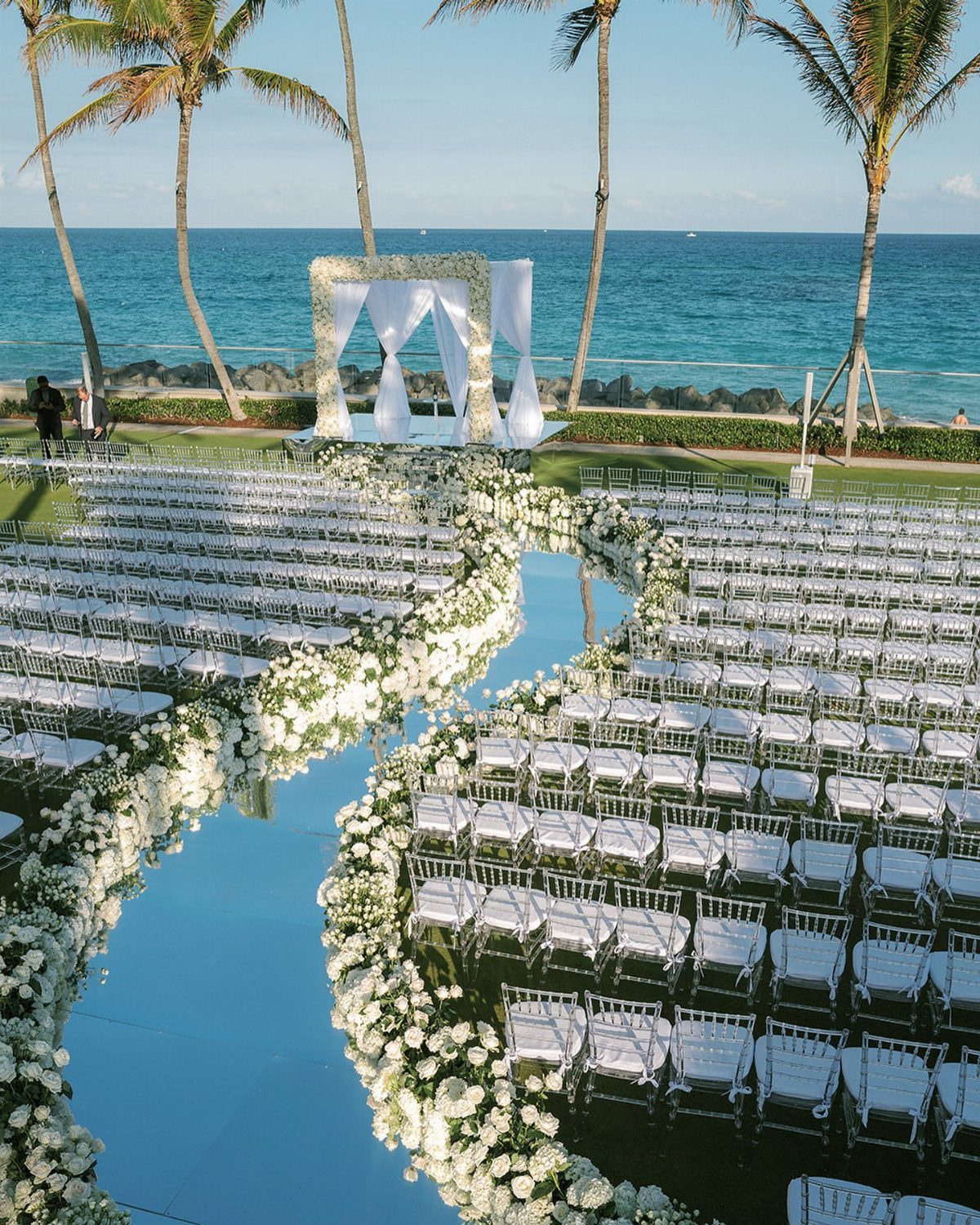 Beach wedding ceremony setup with white chairs, floral aisle, and ocean backdrop.