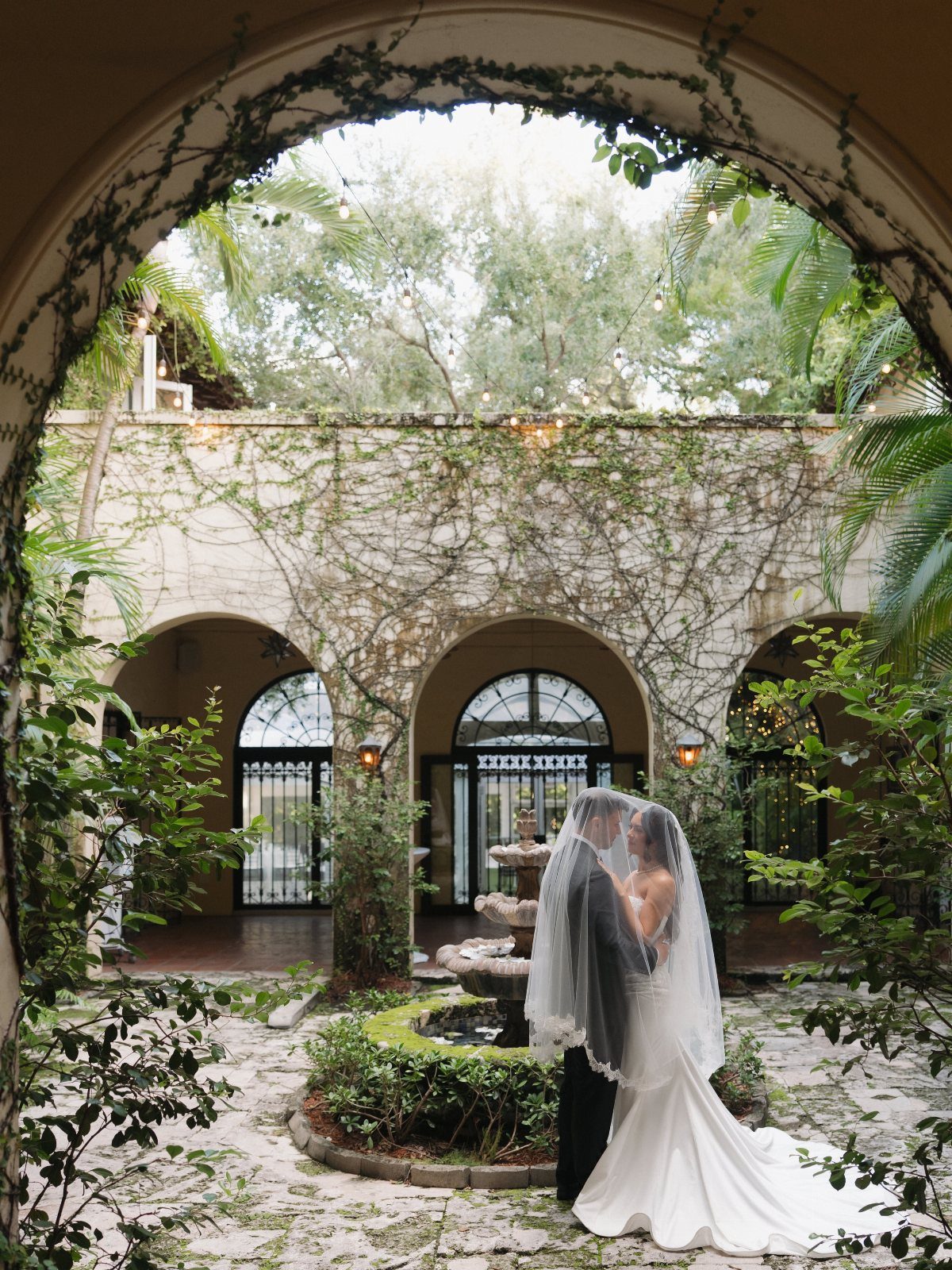 Bride and groom under a veil in a Mediterranean courtyard with stone arches.