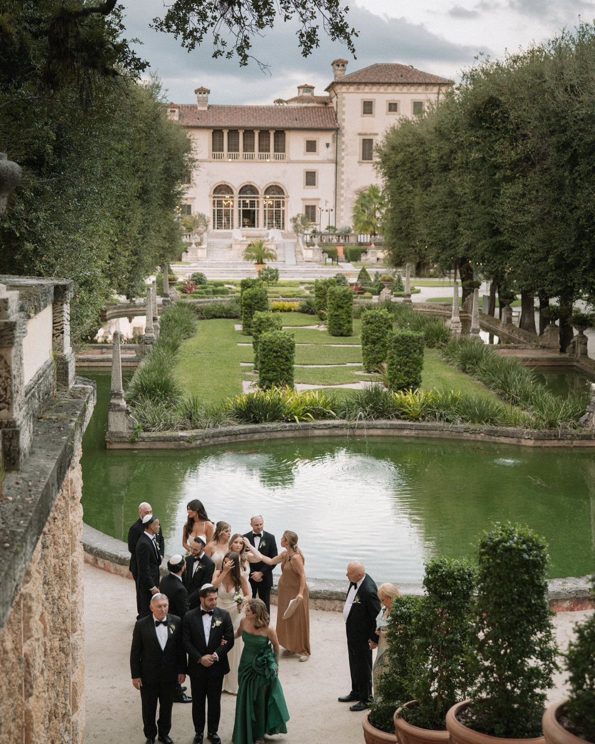 Wedding party in a formal garden with a European-style villa in the background.