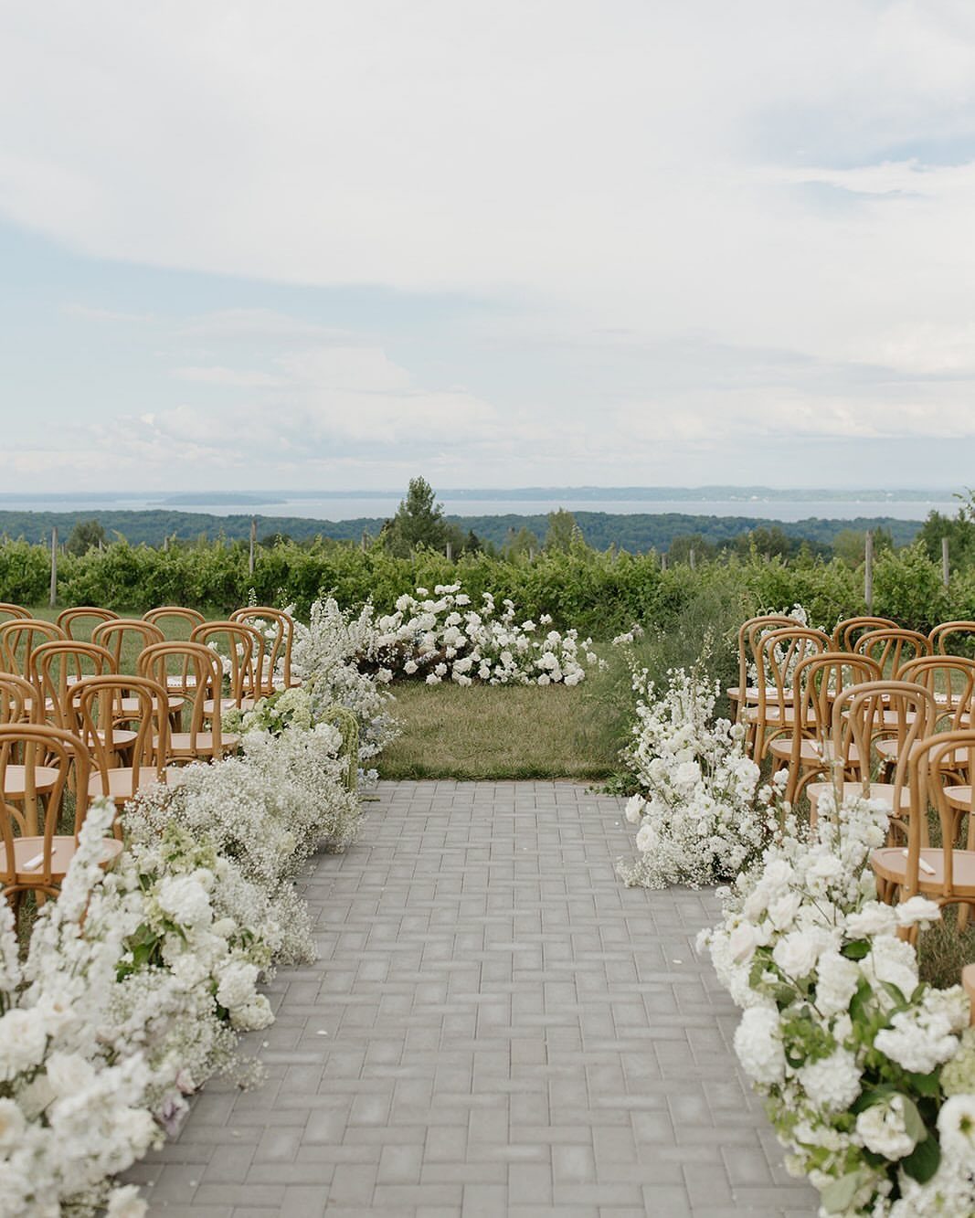 Outdoor wedding ceremony aisle with white flowers and wooden chairs overlooking green vineyards.
