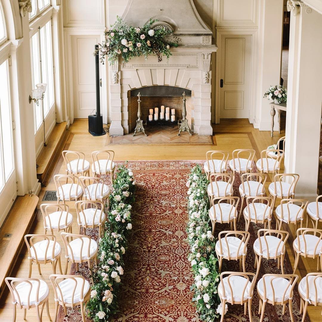 Wedding ceremony setup with white chairs and a floral stone fireplace in a manor.