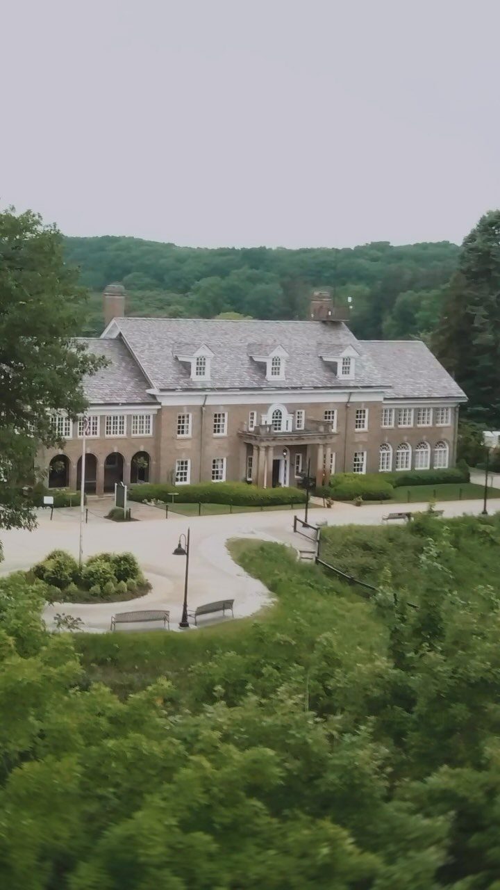 Large brick mansion with grey roof and white trim surrounded by lush green foliage