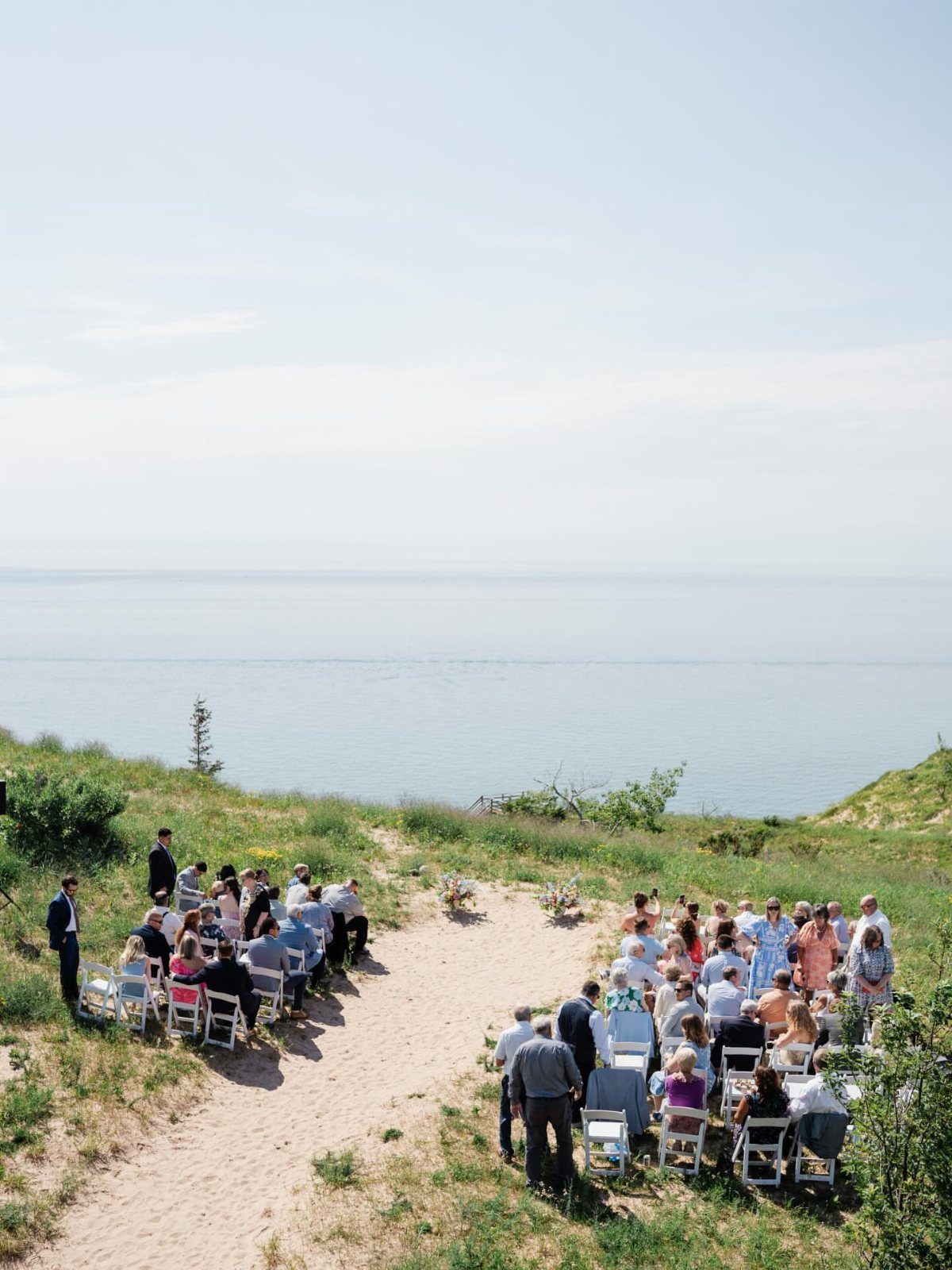 Wedding ceremony guests seated on a grassy bluff overlooking a vast blue lake.