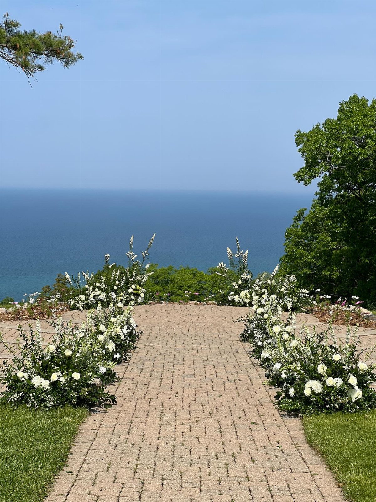 Brick pathway with white flowers leading to a scenic blue water overlook.