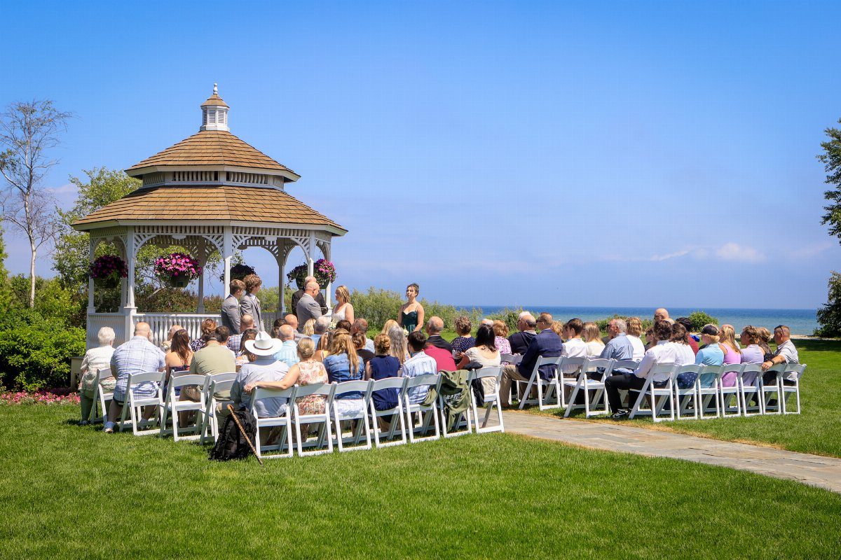 Wedding ceremony with guests in white chairs facing a gazebo by a blue lake.