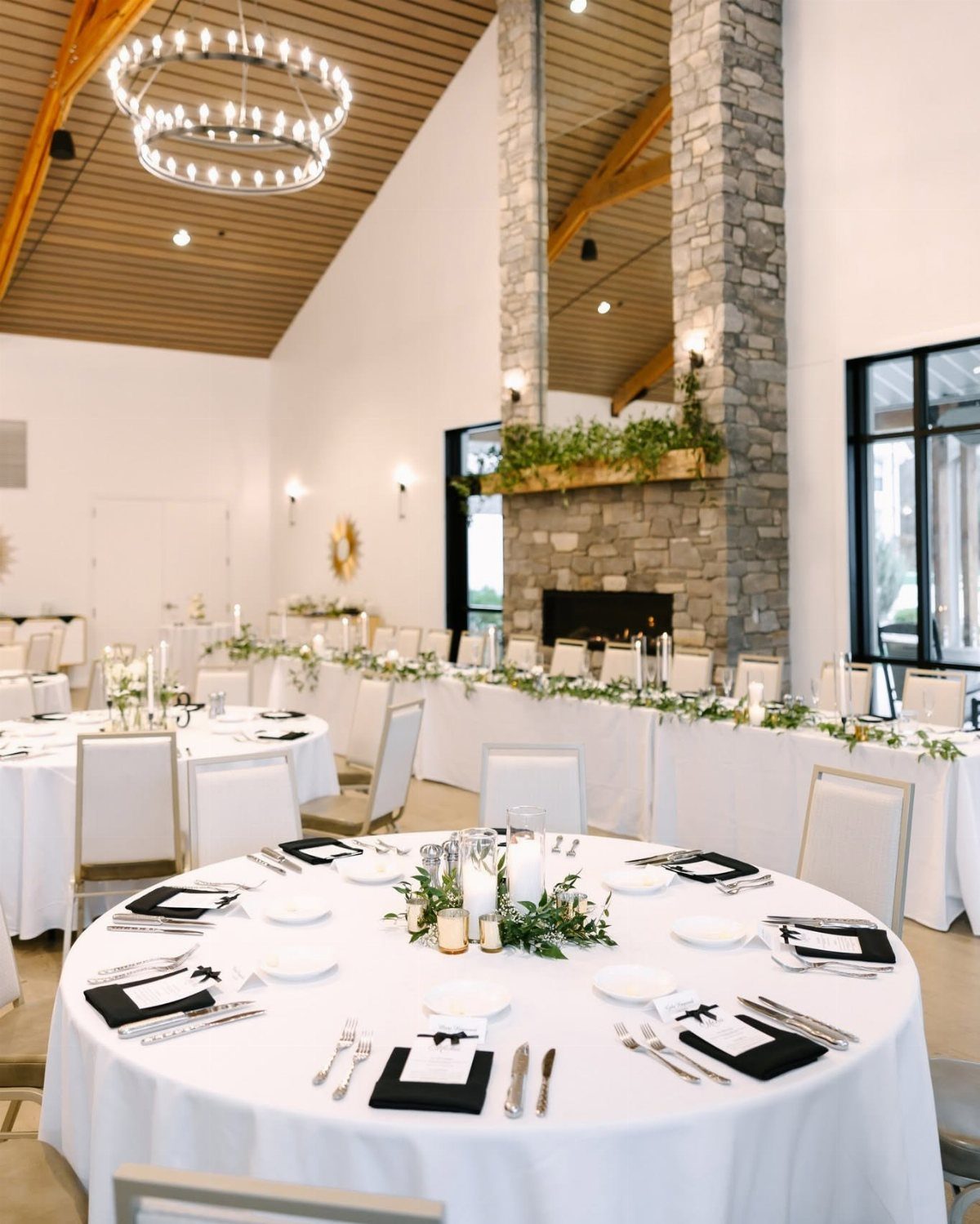 Wedding reception room with white clothed tables, greenery, and a stone fireplace.