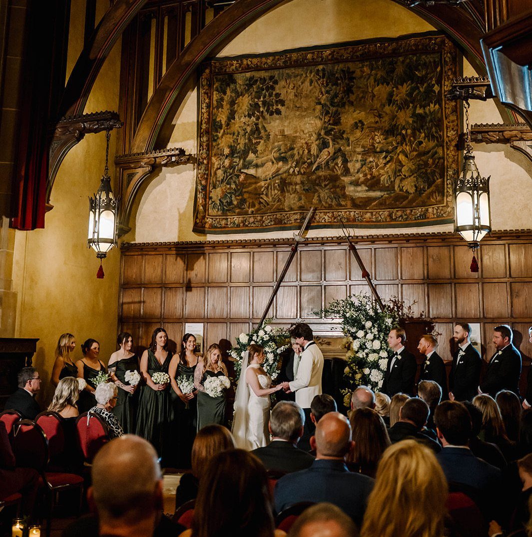 Wedding ceremony in a Tudor-revival mansion hall with wood paneling and a large tapestry.