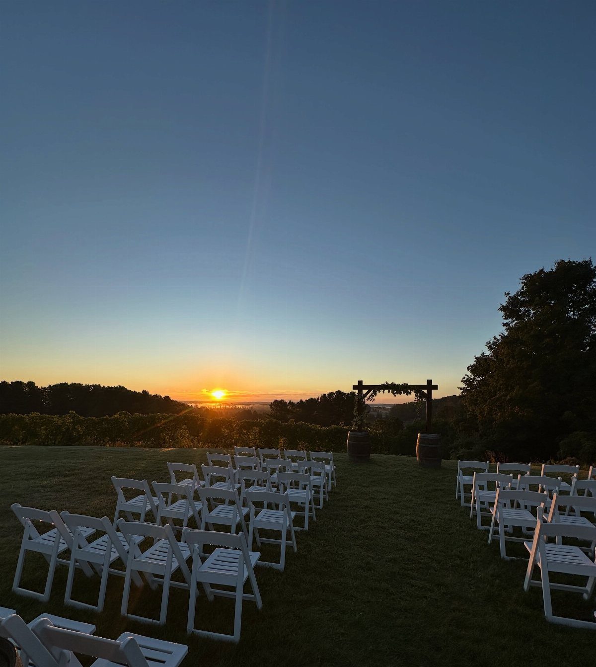 Outdoor wedding ceremony setup with white chairs and a wooden arbor at sunset.