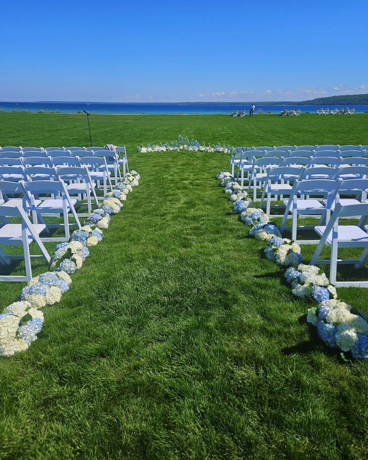 Outdoor wedding ceremony setup with white chairs and blue hydrangeas overlooking a blue lake.