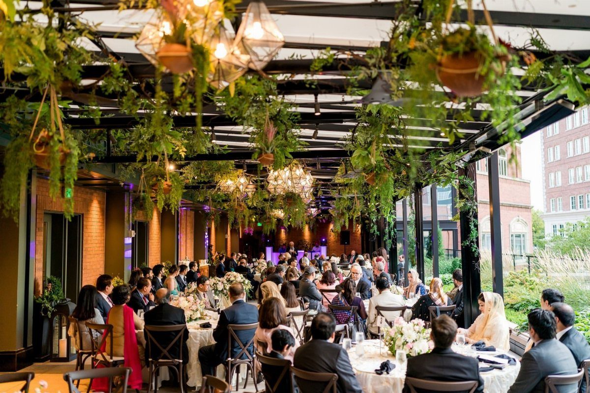 Outdoor wedding reception with guests at round tables under a plant-covered pergola.