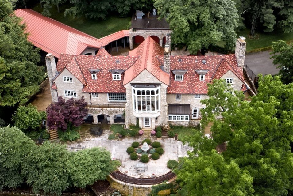 Large stone estate with red roof and circular courtyard fountain surrounded by green trees.