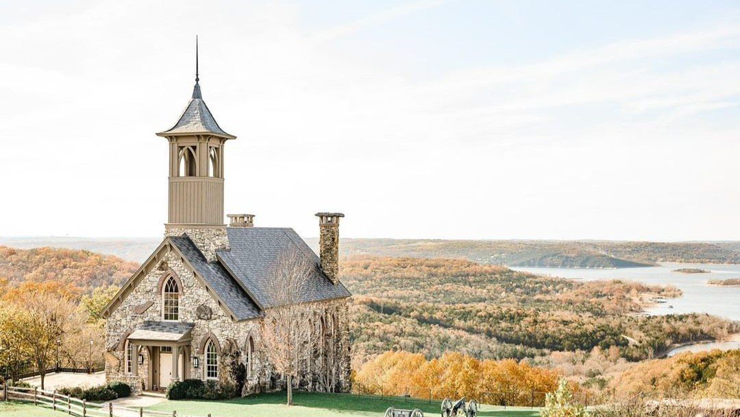 Stone chapel with a steeple overlooking a scenic autumn landscape and lake.