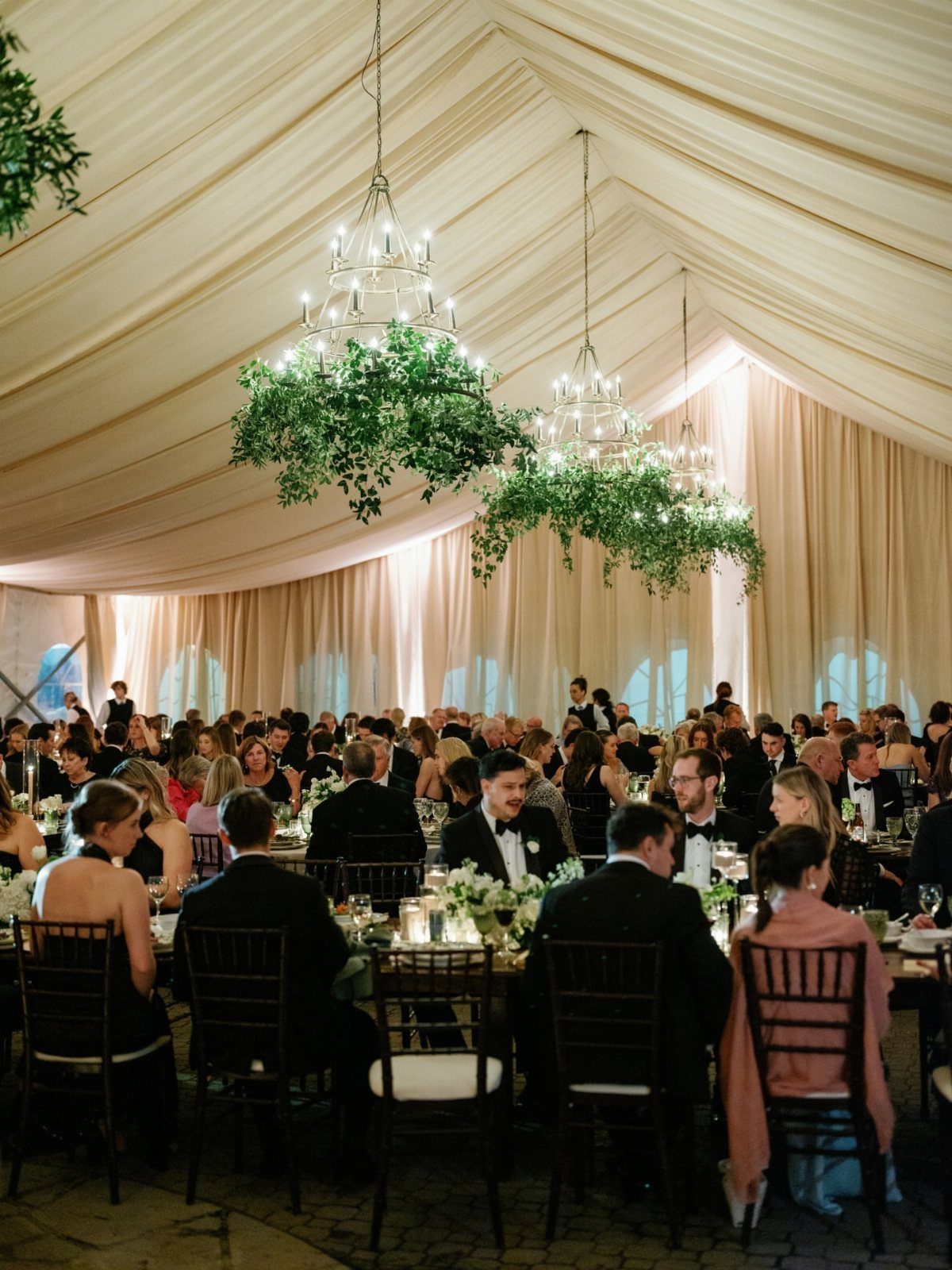 Wedding guests dining under white tent draping with greenery-covered chandeliers above.