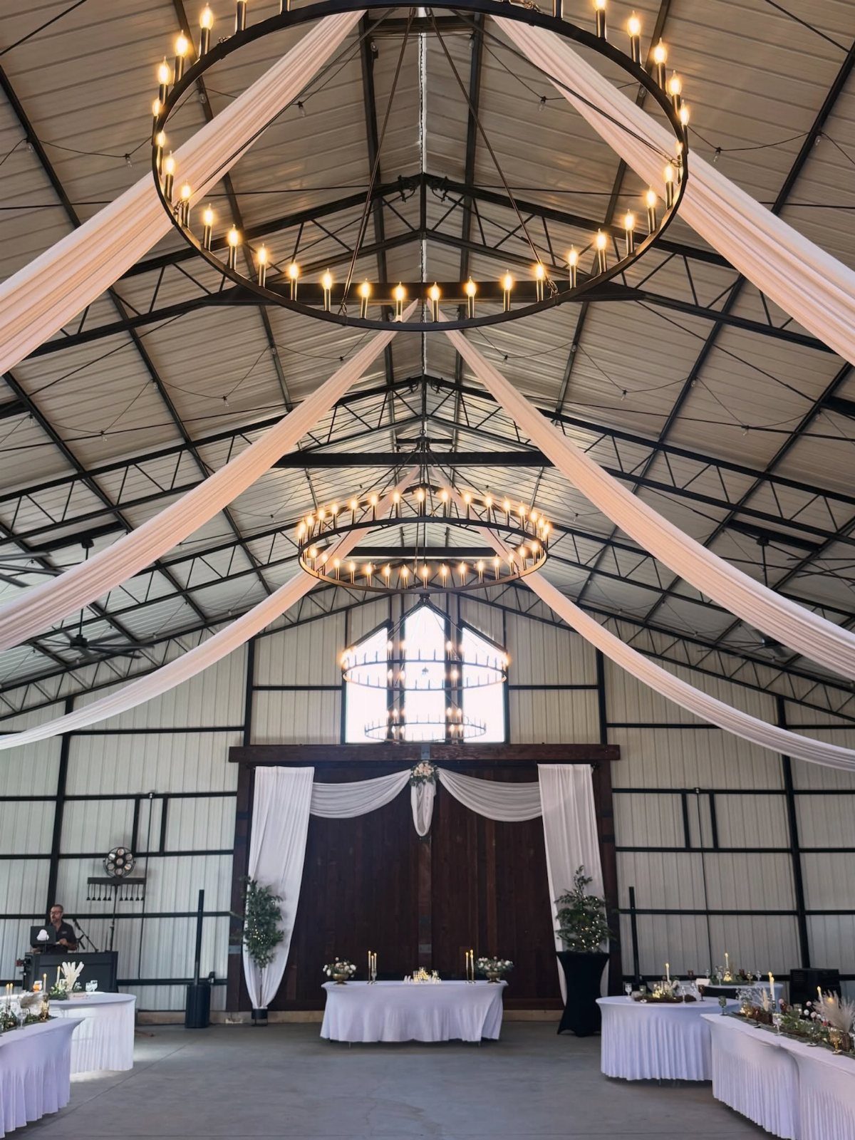 Barn wedding venue interior with white fabric draping and large circular chandeliers.